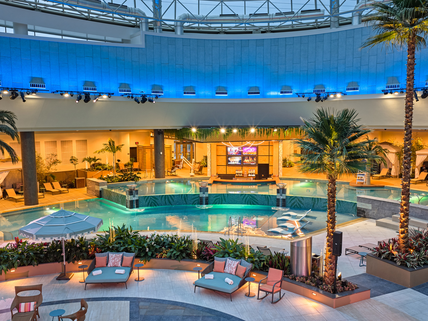 Indoor pool area with lounge chairs, palm trees, ambient lighting, and poolside cabanas under a high ceiling.