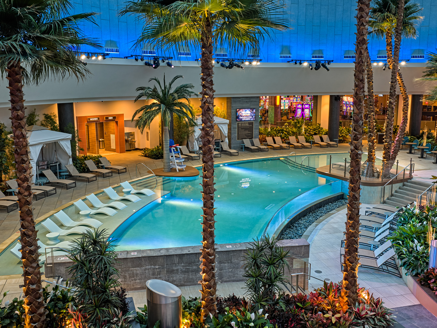 Indoor pool area with lounge chairs, palm trees, cabanas, and lush plants under blue ceiling lights; lifeguard chair and restrooms visible in the background.