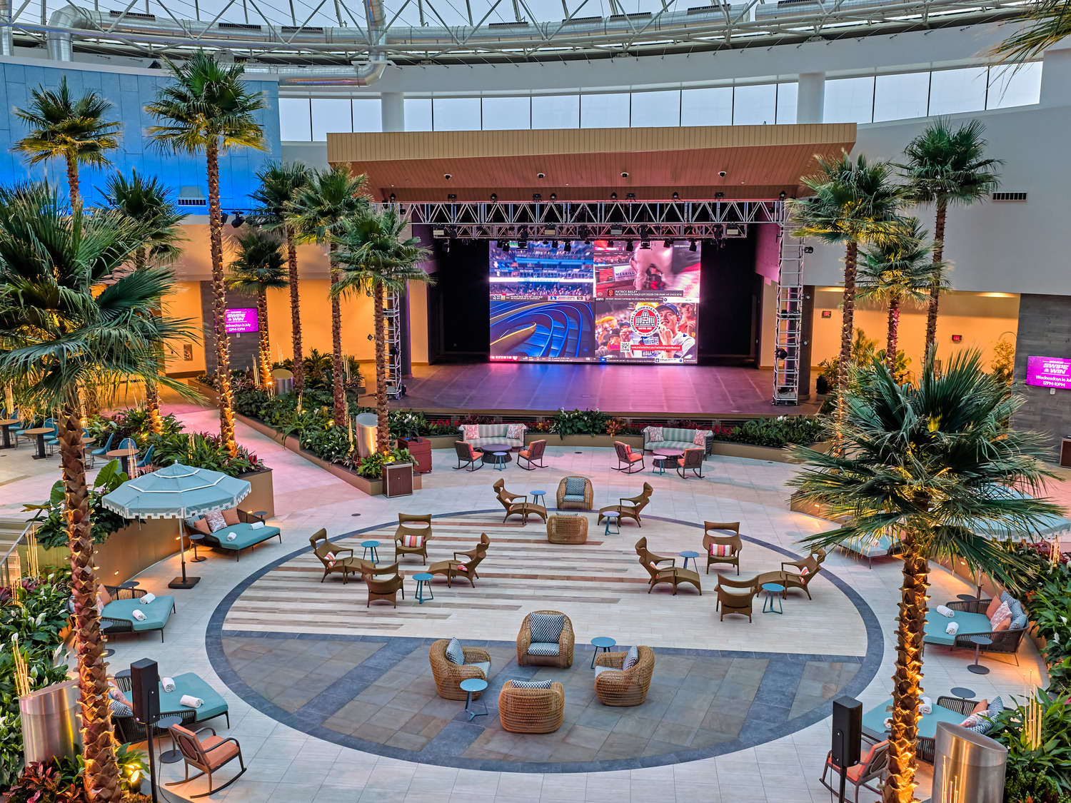 Spacious indoor lounge area with wicker seating, palm trees, and a large stage featuring a bright screen under a high glass ceiling.