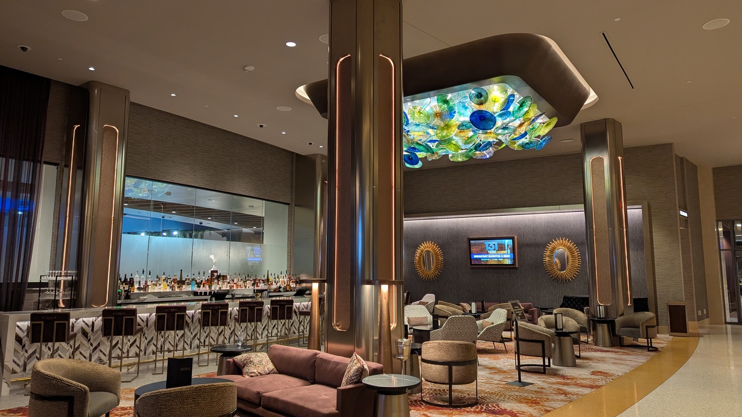 Modern hotel lounge with a bar, contemporary seating, decorative columns, and a colorful glass ceiling sculpture.