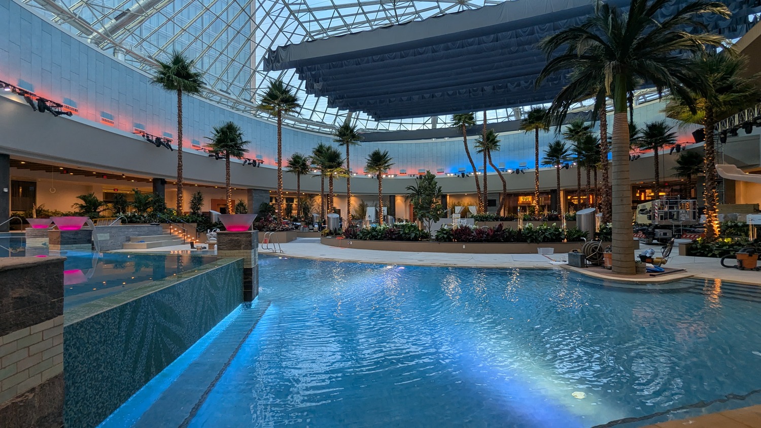 Indoor swimming pool with blue water, surrounded by palm trees and plants, under a large glass dome with artificial lighting and modern architecture.