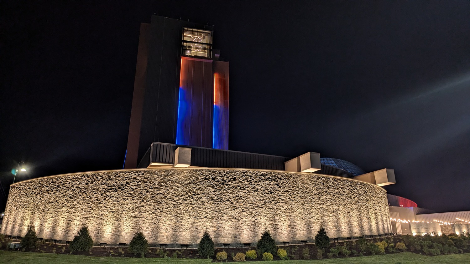 A modern, circular building with a tall rectangular tower is illuminated at night by white and colored lights, with landscaped shrubs in front.