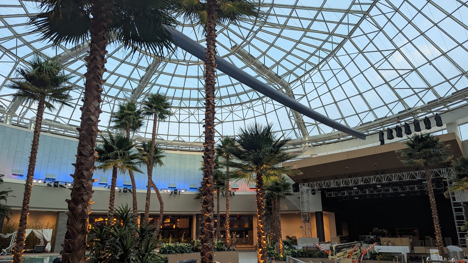 Indoor atrium with tall palm trees, a glass dome ceiling, construction materials, and a stage setup, suggesting ongoing preparations or renovations.