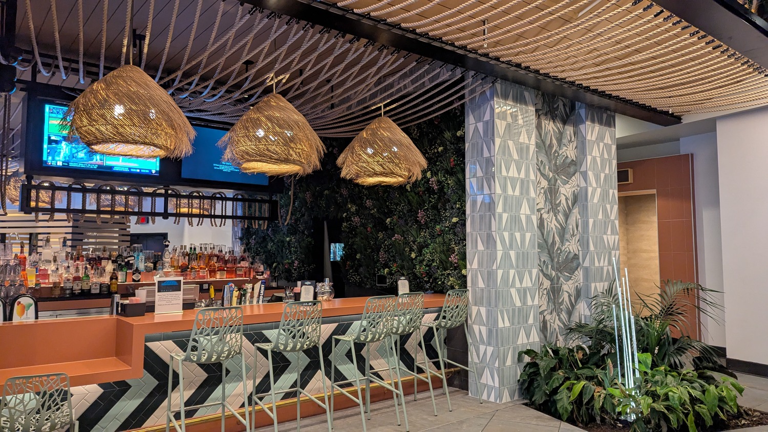 A modern bar with geometric patterned counter, hanging wicker lamps, high chairs, a TV above, and decorative plants on the wall and floor.