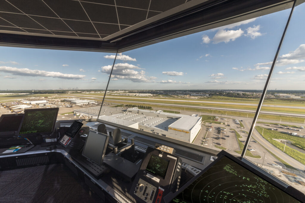 View from an airport control tower overlooking runways, terminals, and aircraft, highlighting modern airport design elements, with radar screens and control equipment in the foreground.