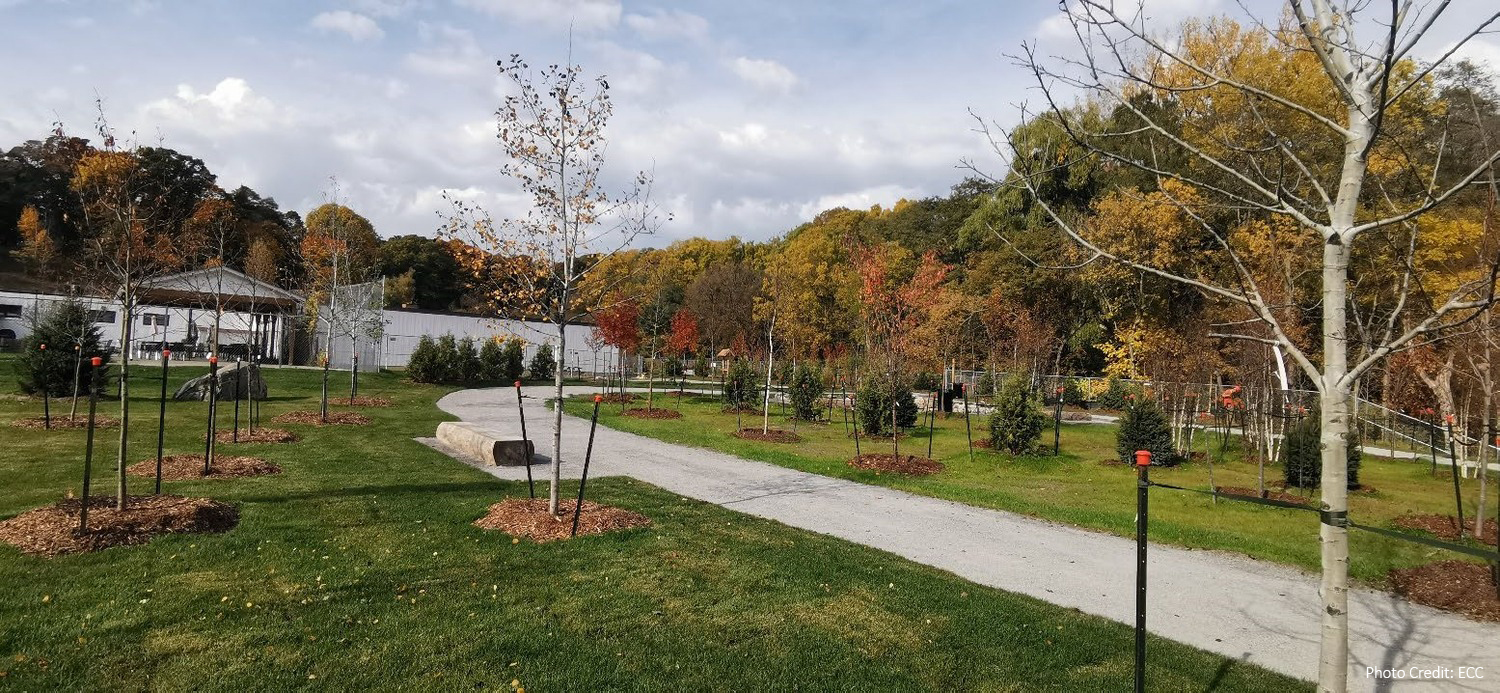 A paved pathway curves through a landscaped park with young trees, grassy areas, and a gazebo in the background under a partly cloudy sky.
