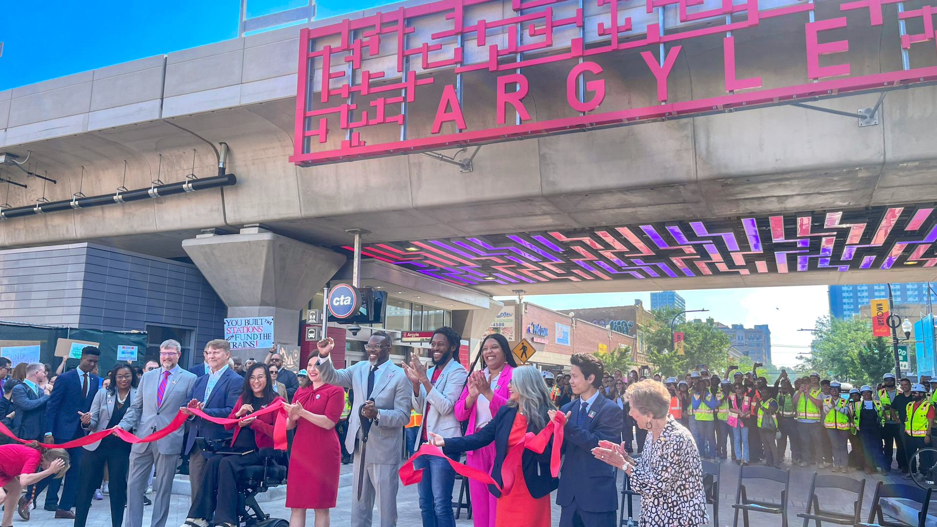 A group of people participates in a ribbon-cutting ceremony beneath a large red "Argyle" sign, celebrating the Red and Purple Modernization Phase One Project, with onlookers and workers in safety vests in the background.