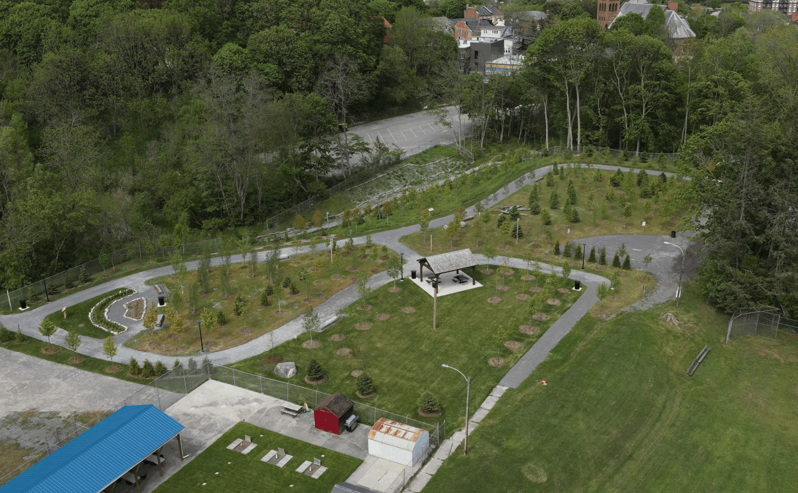 Aerial view of a landscaped park with winding paths, small trees, a shelter, and adjacent parking lot, surrounded by dense greenery and some buildings in the background.
