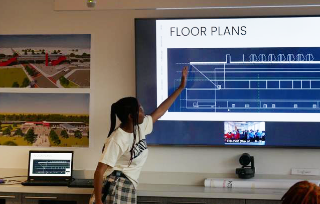 A person points to a floor plan diagram on a large screen during a presentation in a room with architectural images on the wall.
