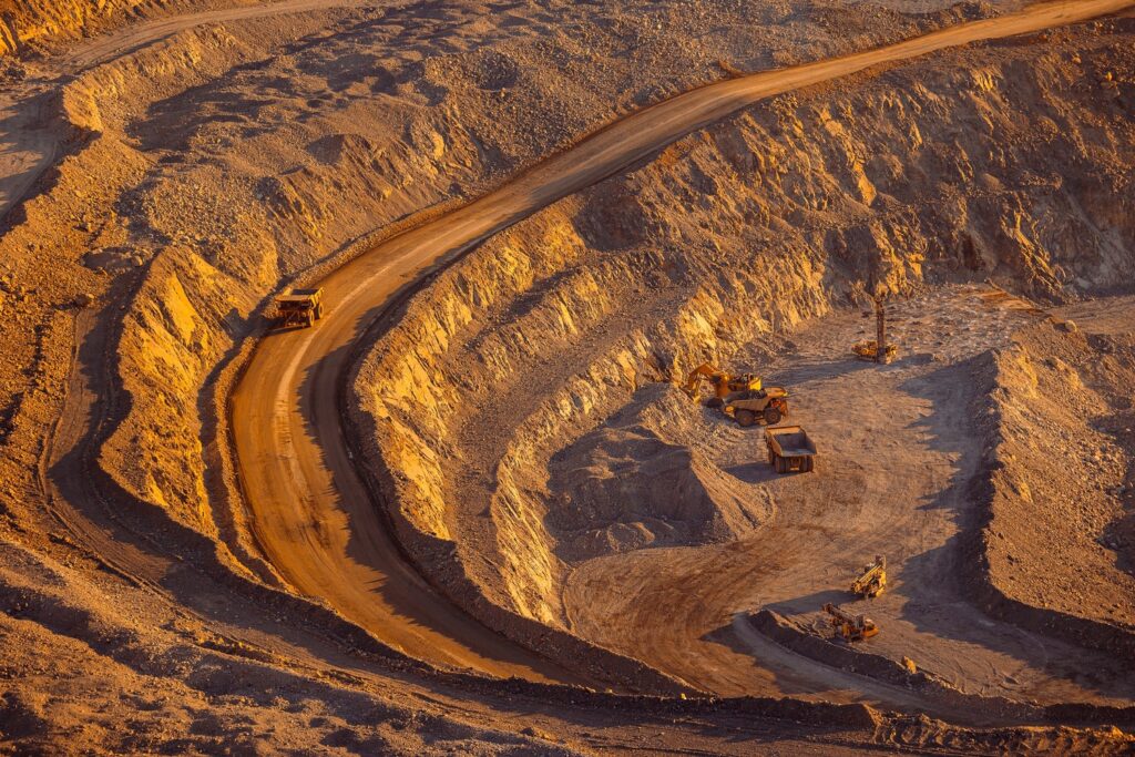 Aerial view of an open-pit mine showing large mining trucks on a winding dirt road and excavation equipment working on terraced slopes.