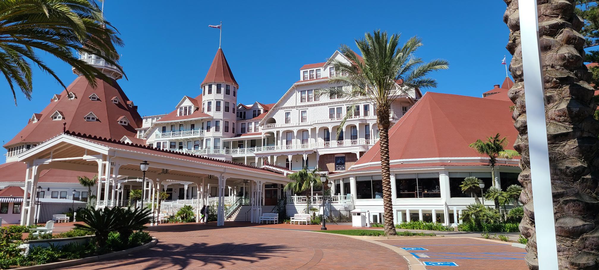 A historic beachfront hotel with white wooden walls, red roofs, palm trees, and clear blue sky, featuring multiple peaked towers and a covered walkway in front.