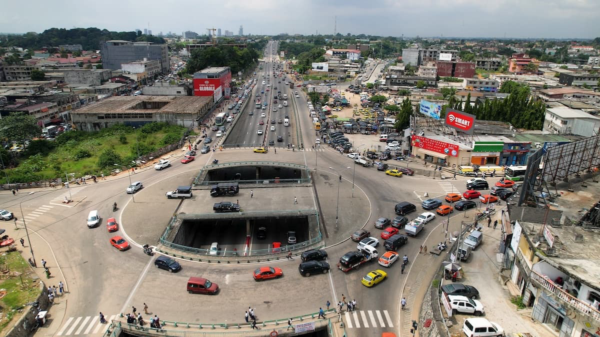 Vue aérienne d'un rond-point urbain très fréquenté par des voitures, des piétons et des magasins, avec un passage souterrain et plusieurs voies de circulation dans un environnement urbain.
