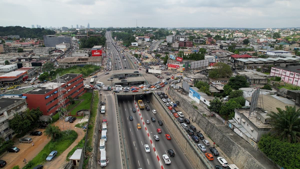 Vue aérienne d'une autoroute à plusieurs voies dans une zone urbaine à forte circulation, entourée de bâtiments, d'arbres et d'un ciel nuageux.
