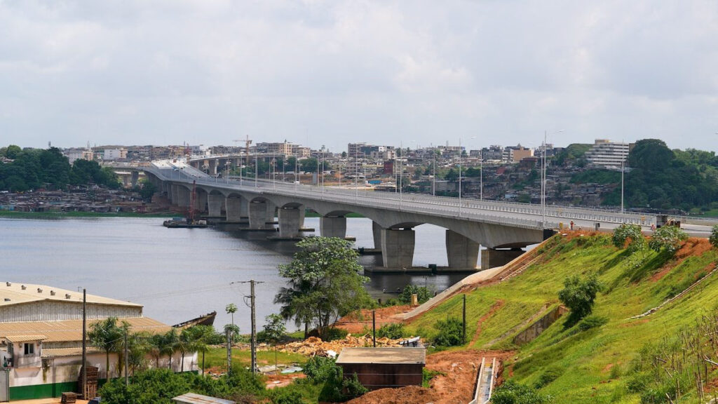 A concrete bridge crosses a wide river, connecting two urban areas with buildings and greenery visible on both sides under a cloudy sky.