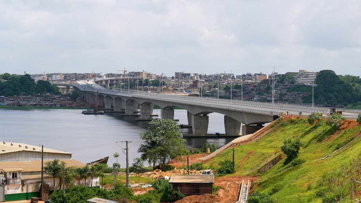 Un grand pont en béton enjambe une large rivière, reliant deux zones urbaines dont les bâtiments sont visibles de part et d'autre sous un ciel nuageux.