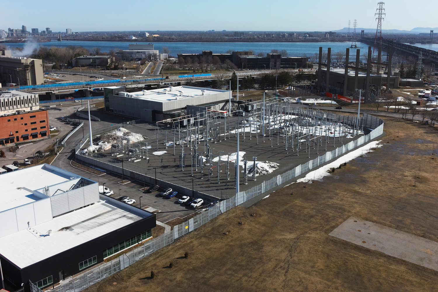 Une sous-station électrique clôturée avec de nombreux poteaux et lignes électriques est située près d'une zone industrielle et d'une rivière, avec des bâtiments, un pont et des voitures garées visibles à proximité.