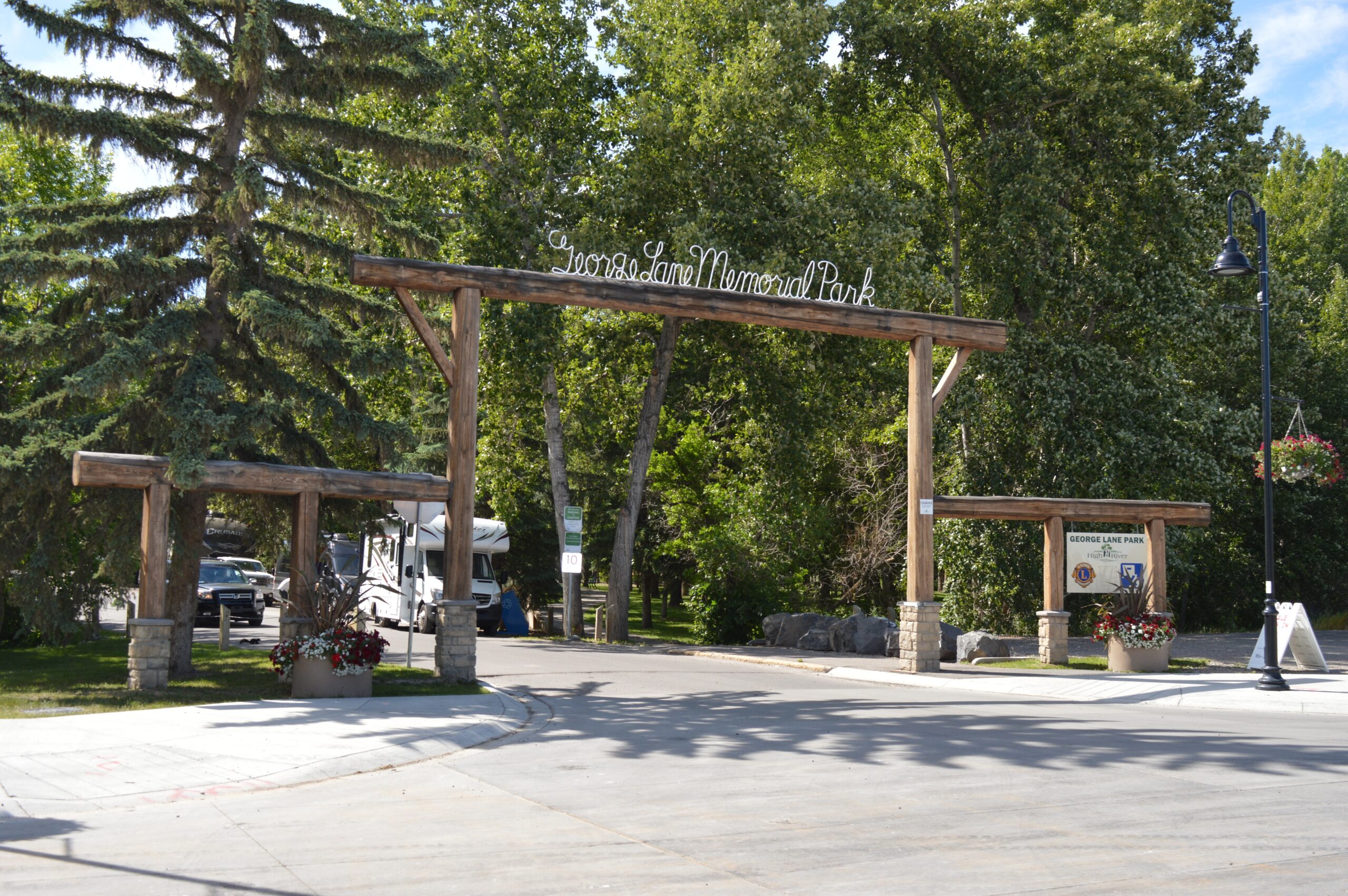 Wooden entrance archway with a sign reading "Sandy Lane Memorial Park," surrounded by trees, flower planters, and informational boards on a sunny day.