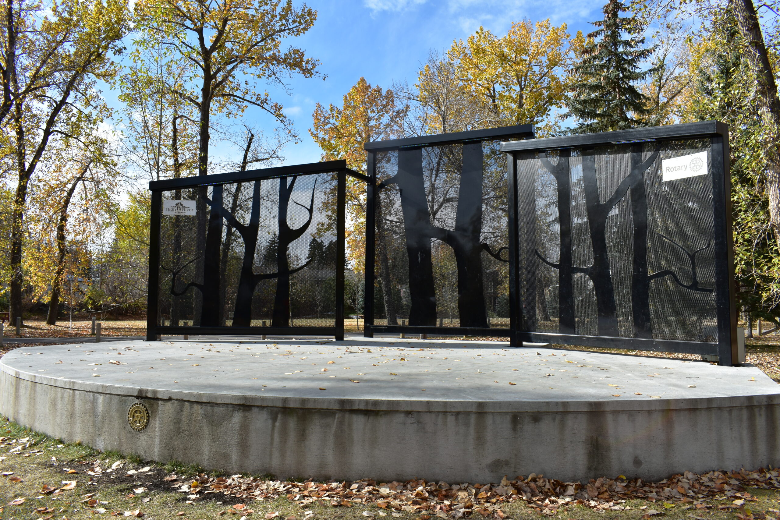 A concrete platform with three glass panels featuring etched tree designs stands in a park surrounded by autumn trees and fallen leaves.