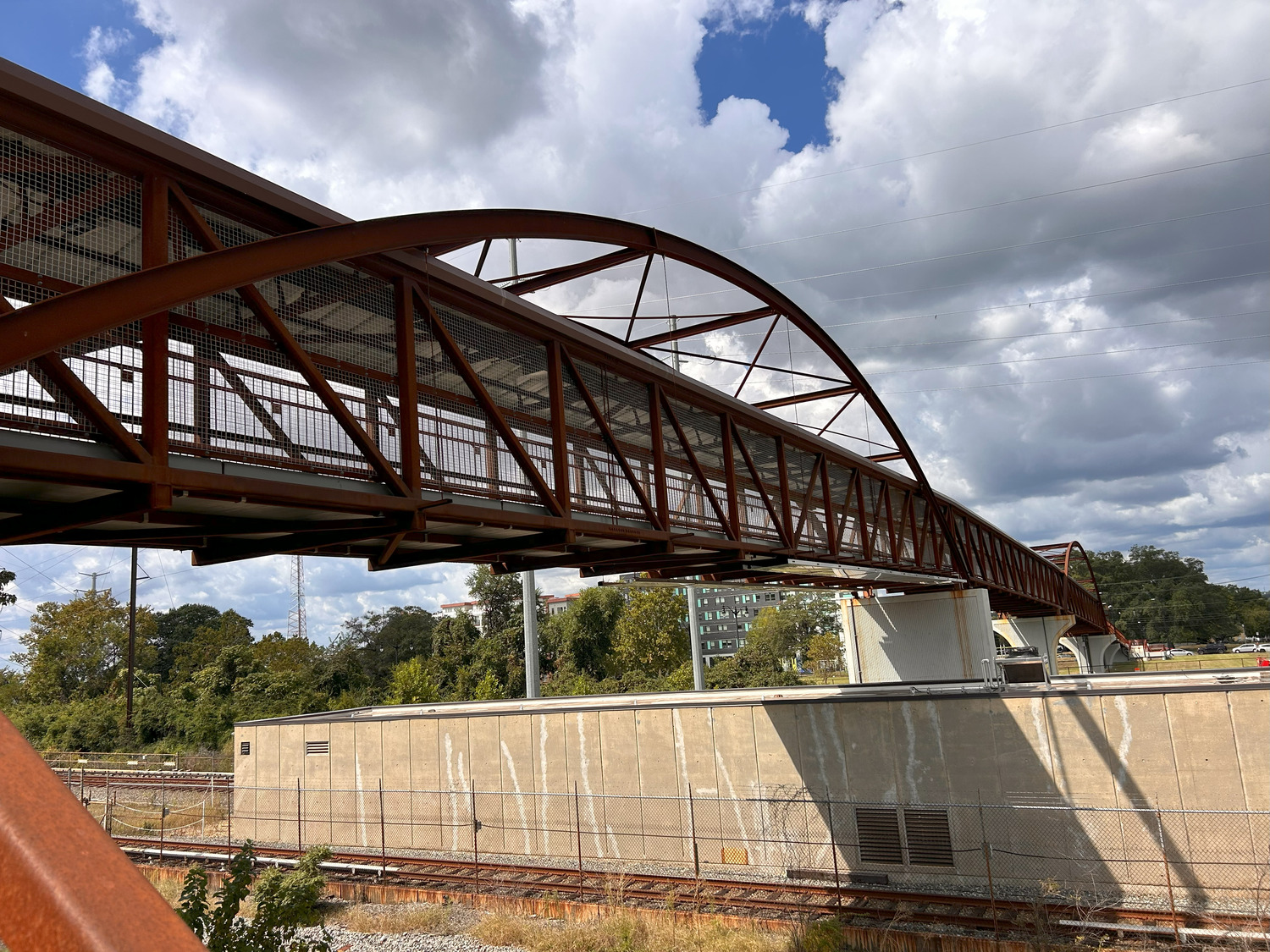 Un pont piétonnier en métal rouge enjambe des voies ferrées et un mur en béton, avec des arbres et des bâtiments visibles à l'arrière-plan sous un ciel nuageux.