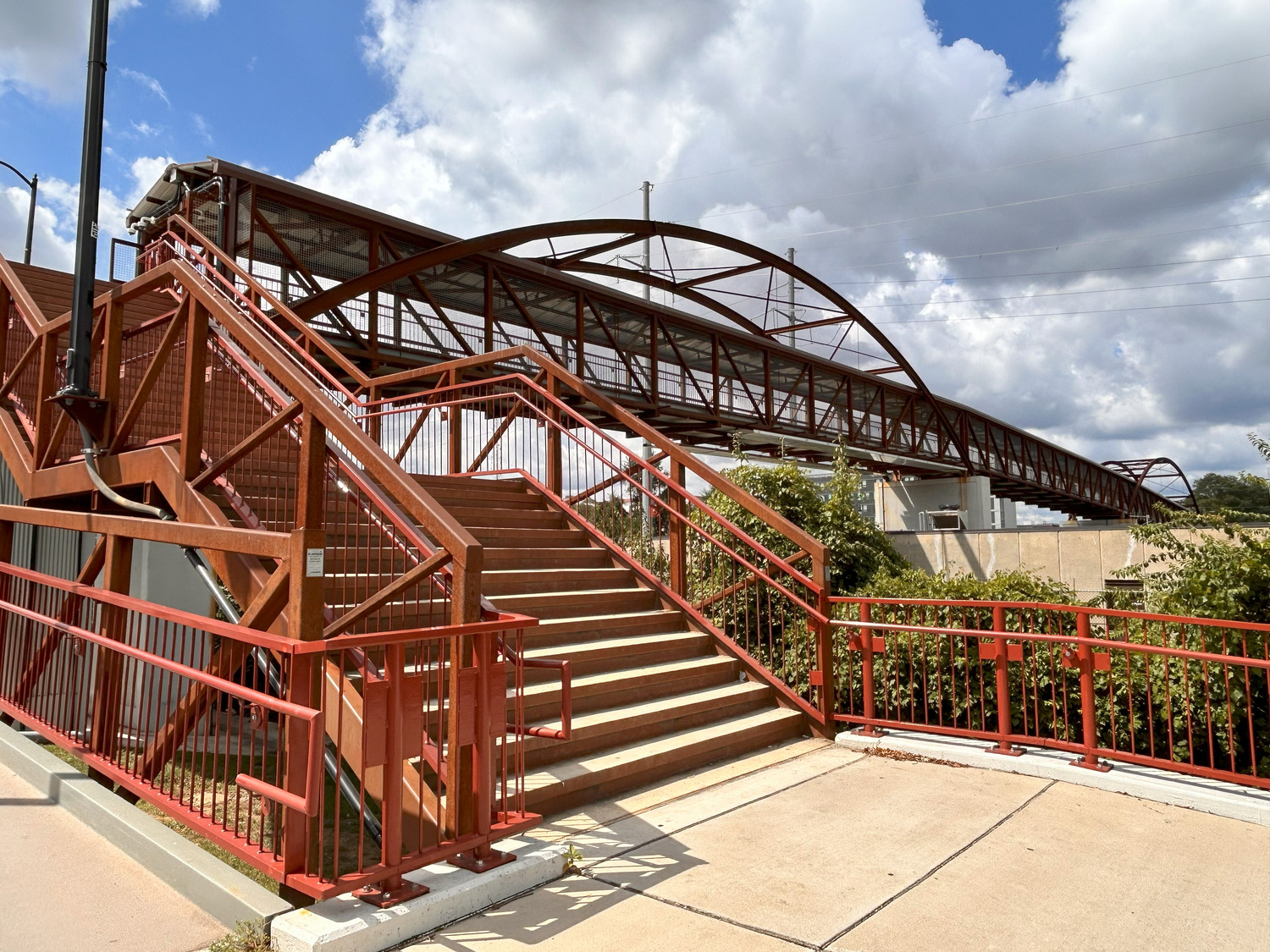 A red metal pedestrian bridge and stairs extend over a roadway, set against a partly cloudy sky with greenery visible around the structure.