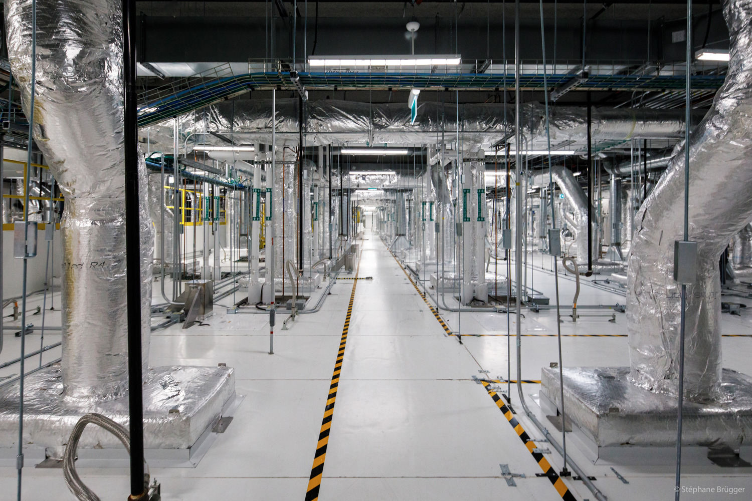 A large industrial room with metallic pipes, ducts, and equipment covered in silver insulation lining both sides of a wide walkway with yellow-black hazard markings.