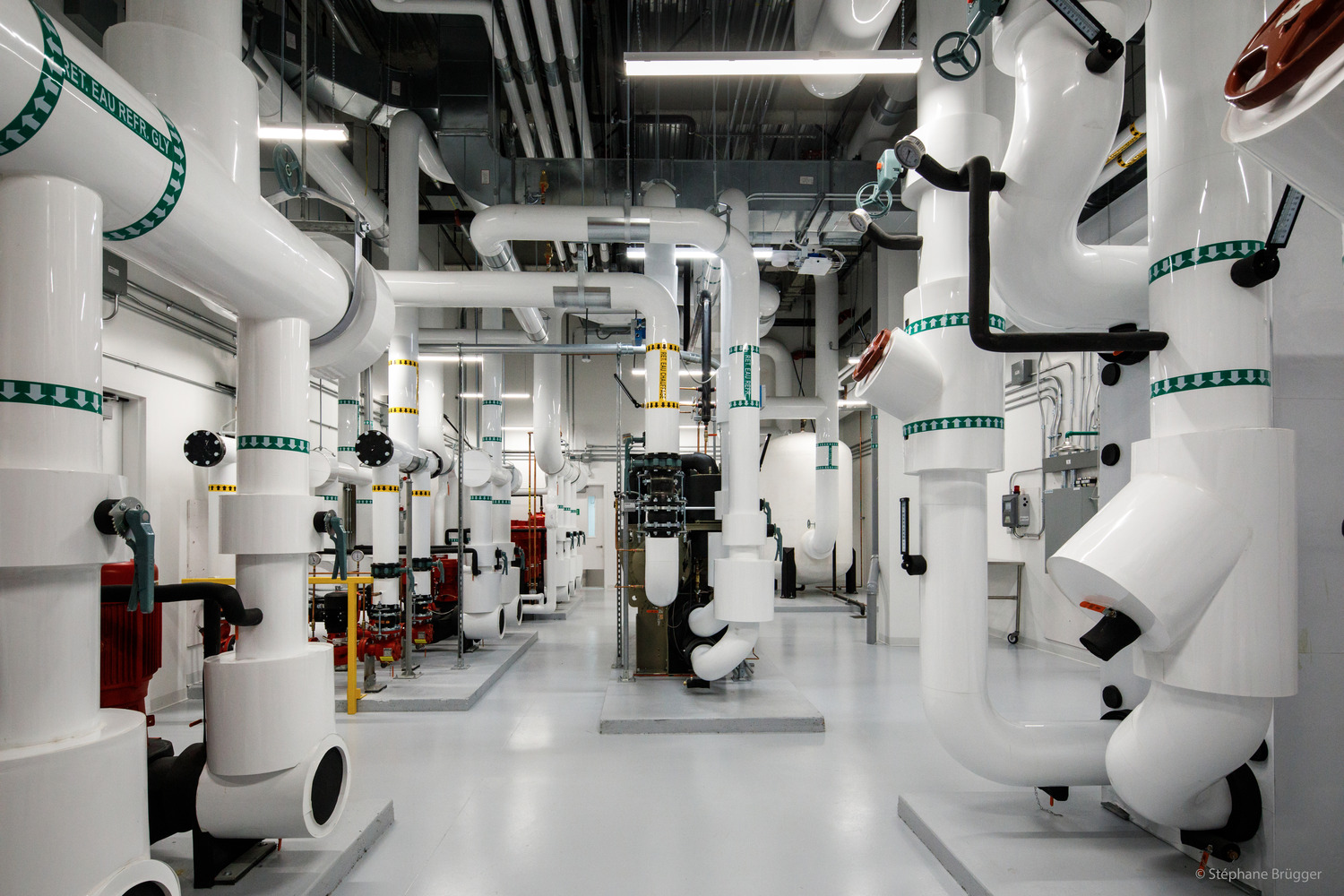 Industrial mechanical room with large white pipes, valves, and equipment distributed throughout a clean, brightly lit space.