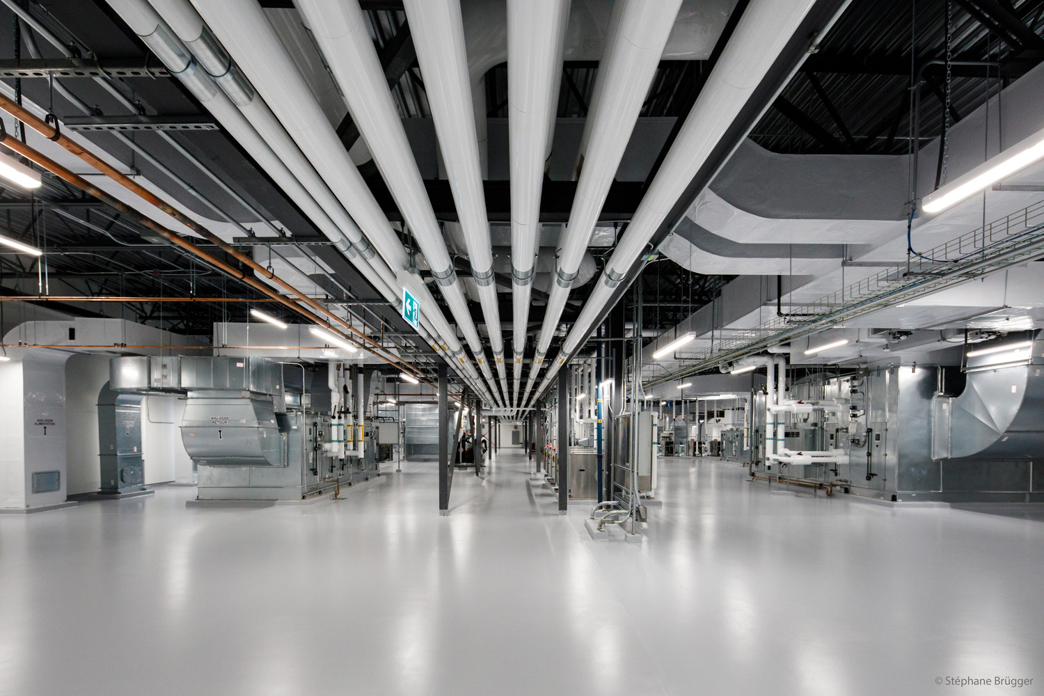 Wide industrial room with shiny floors, exposed pipes and ducts on the ceiling, and large ventilation equipment along the walls.