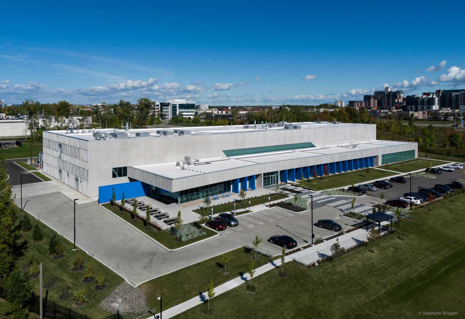 Aerial view of a large, modern, rectangular building with white and blue exterior, surrounded by a parking lot, trees, and landscaped grounds.