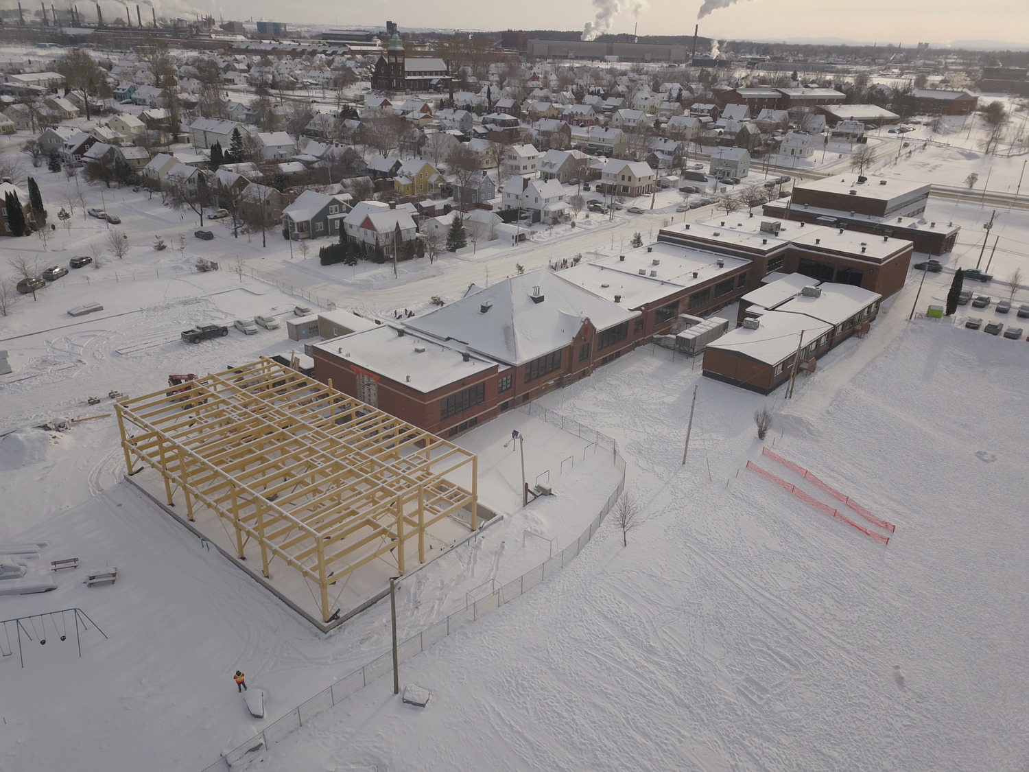 Vue aérienne d'un bâtiment scolaire recouvert de neige et d'une structure en acier en construction à côté, dans un quartier résidentiel.