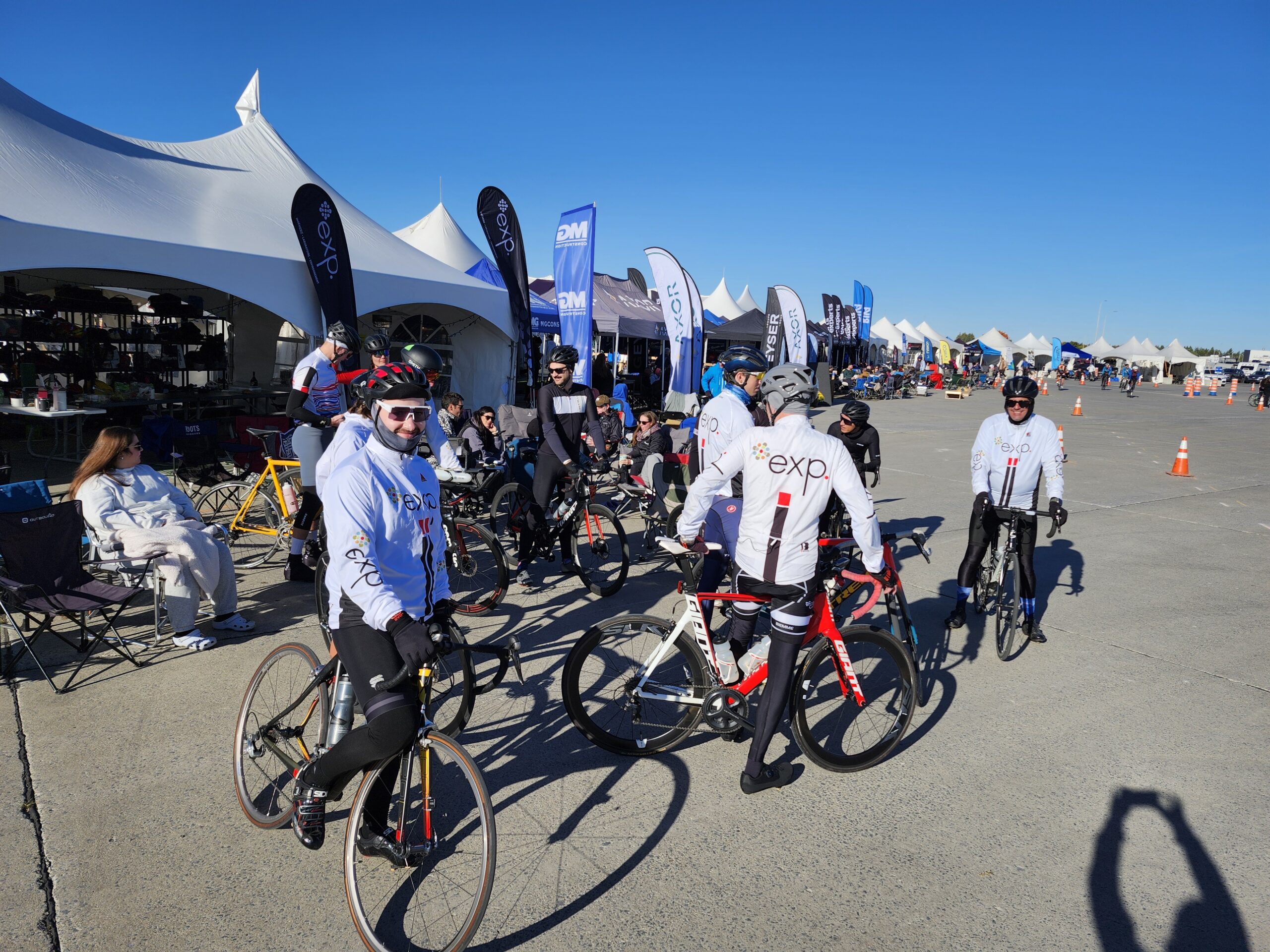 Un groupe de cyclistes vêtus de maillots blancs assortis se rassemble près de tentes lors d'un événement en plein air par une journée ensoleillée.