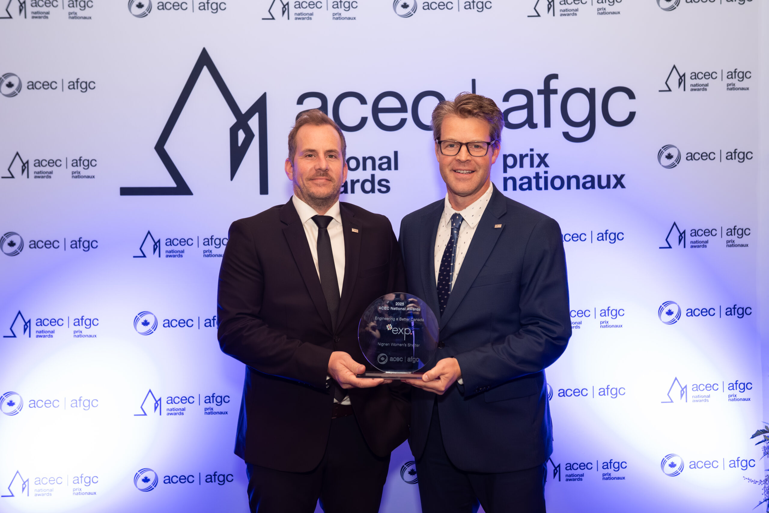 Two men in suits stand in front of an ACEC-ACGC backdrop, holding a glass award plaque together and facing the camera.