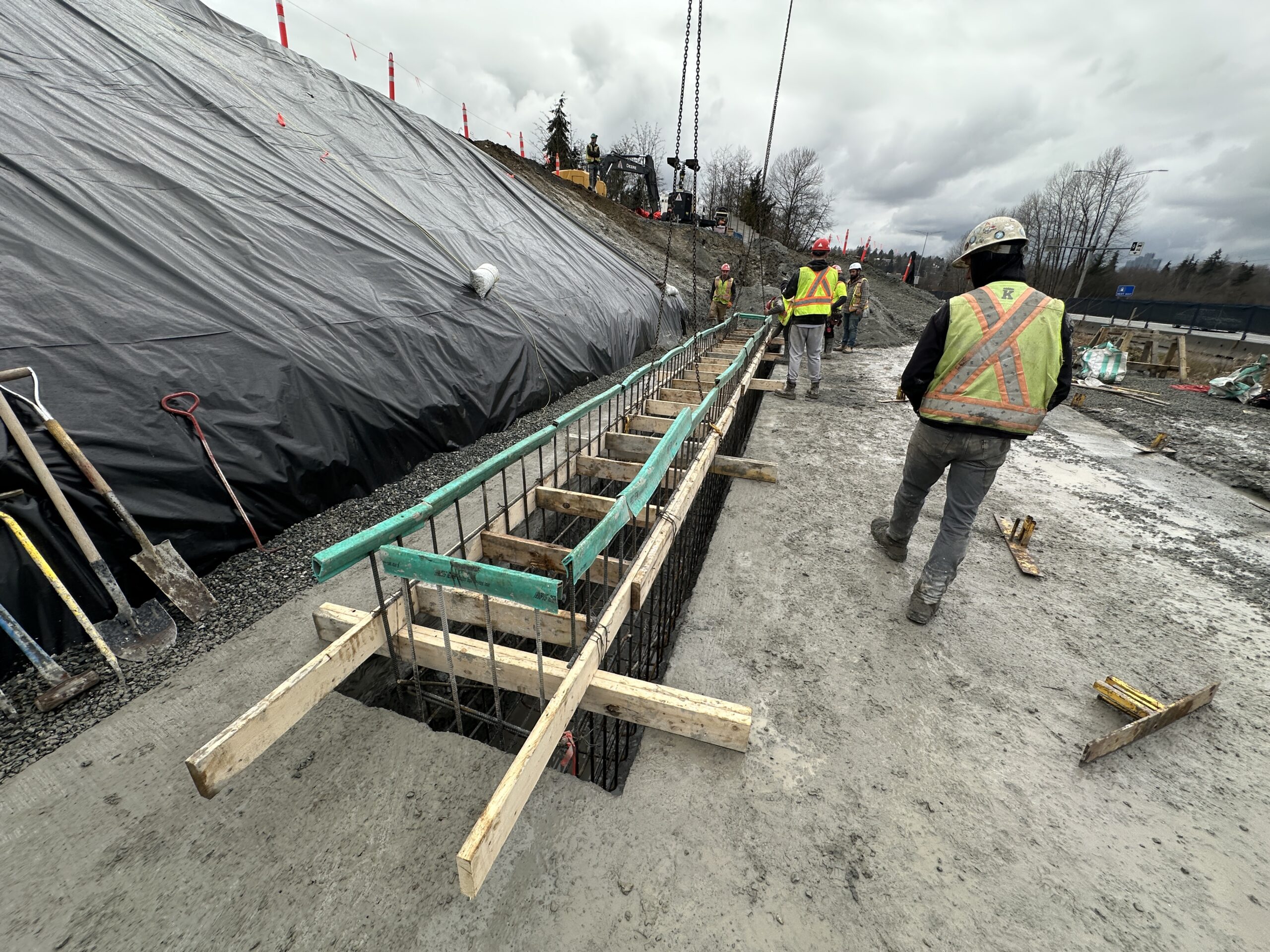 Construction workers walk near rebar and wooden formwork for a concrete structure at a muddy outdoor construction site on a cloudy day.