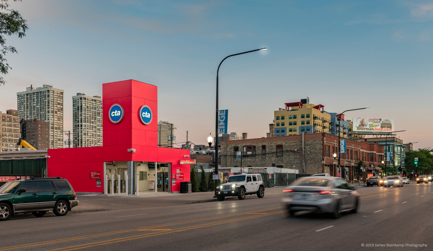 Street view of a red CTA building with vehicles driving by, surrounded by city buildings under a partly cloudy evening sky.