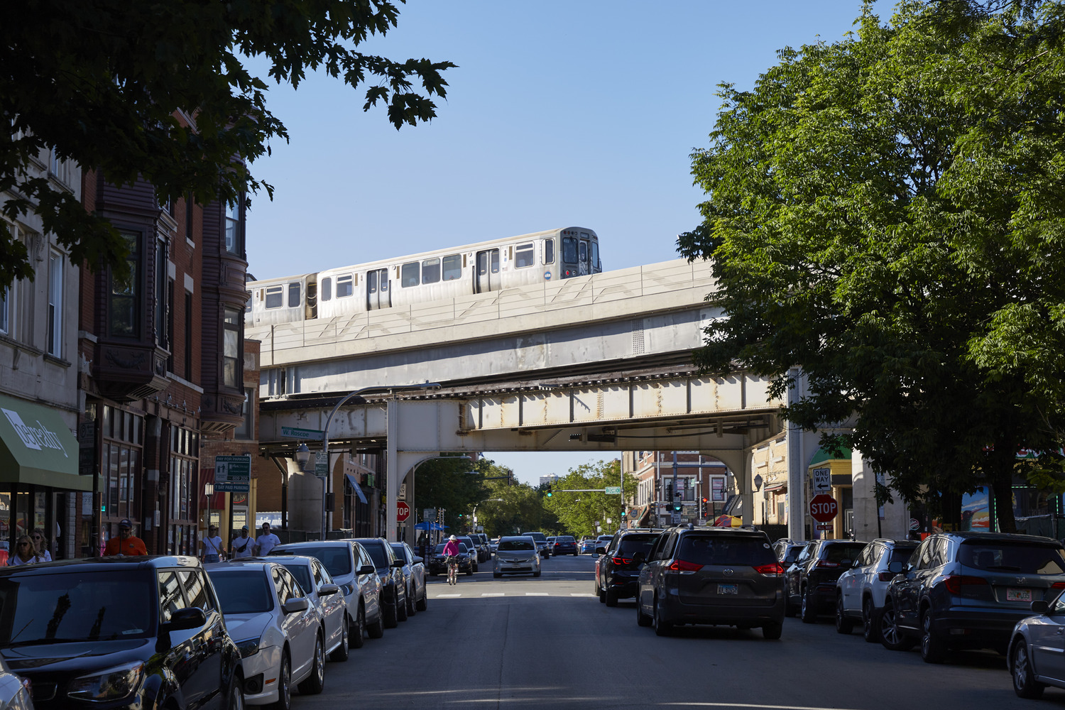A train travels on an elevated track above a busy city street lined with parked cars and trees on a clear day.