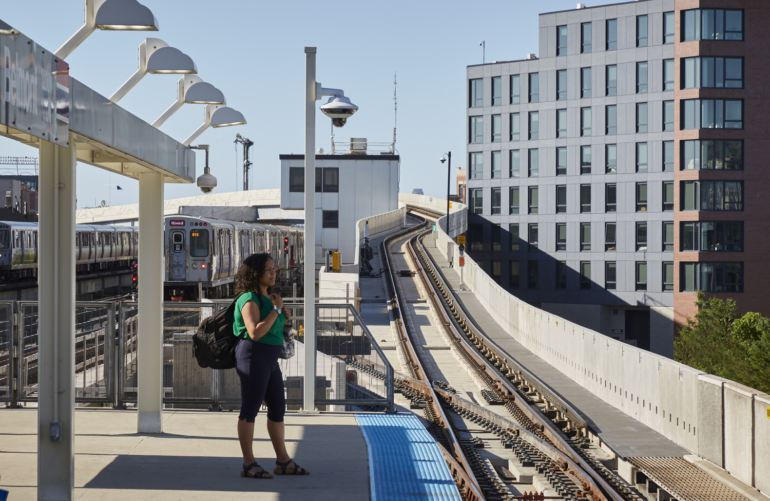 A woman stands on an outdoor train platform next to train tracks, with a train and modern apartment buildings in the background.