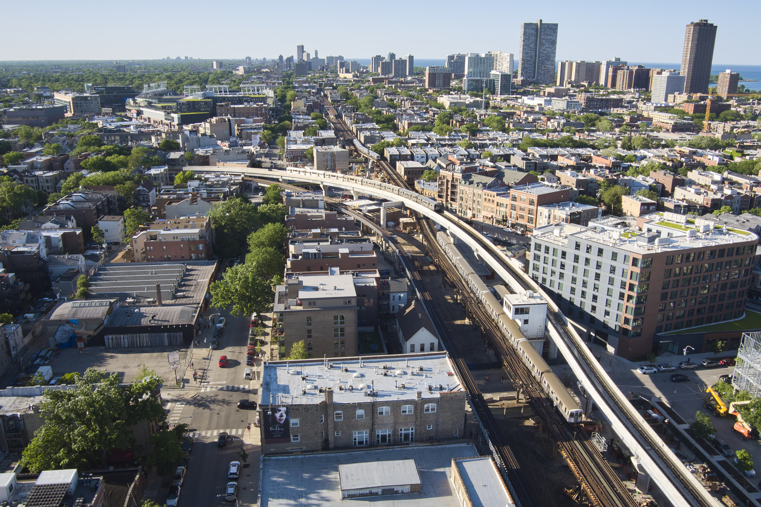 Aerial view of an urban neighborhood with elevated train tracks, residential and commercial buildings, and a city skyline in the distance.