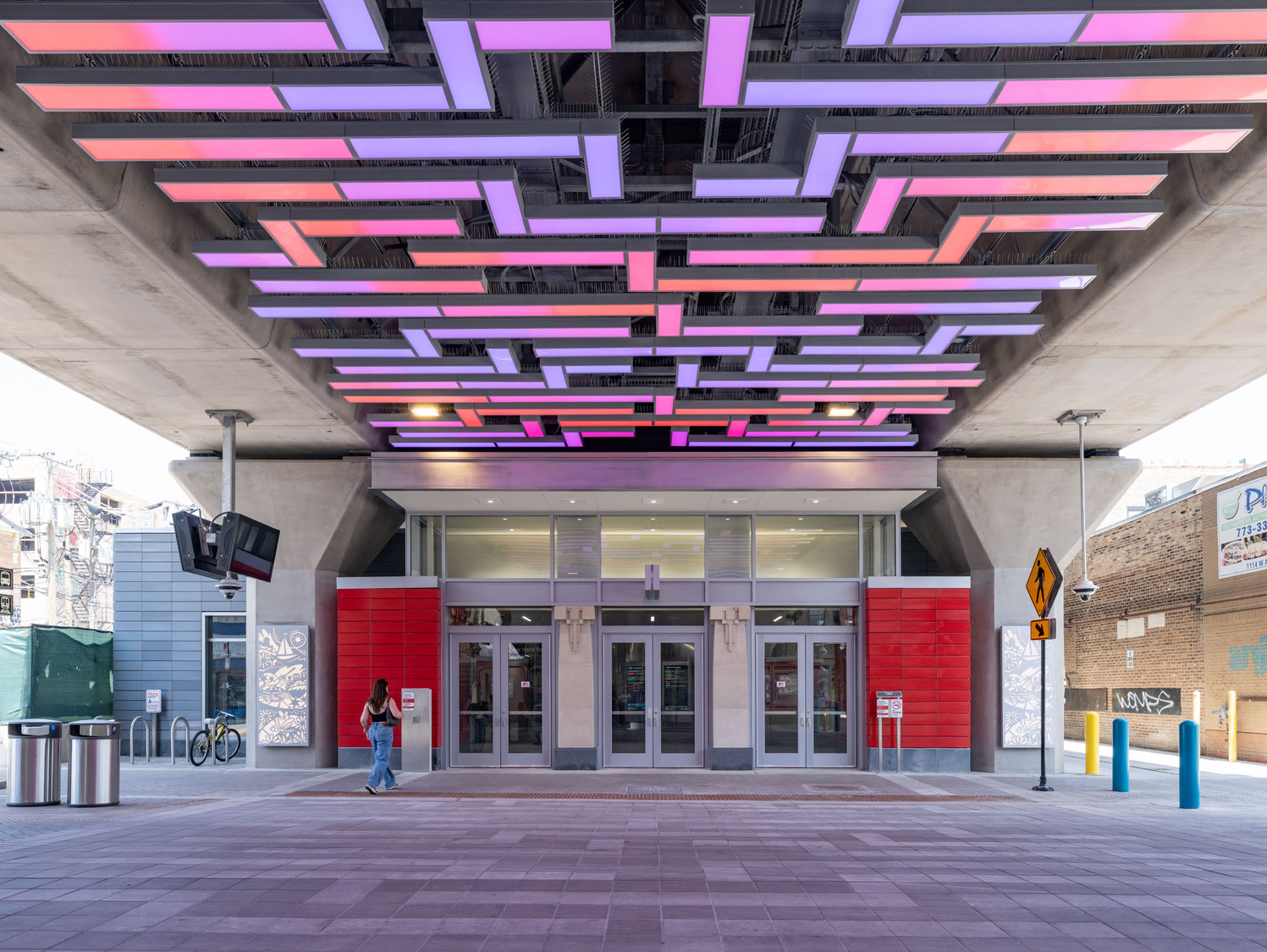 A person walks past the entrance of a modern building under a bridge, with colorful rectangular lights suspended from the ceiling above.