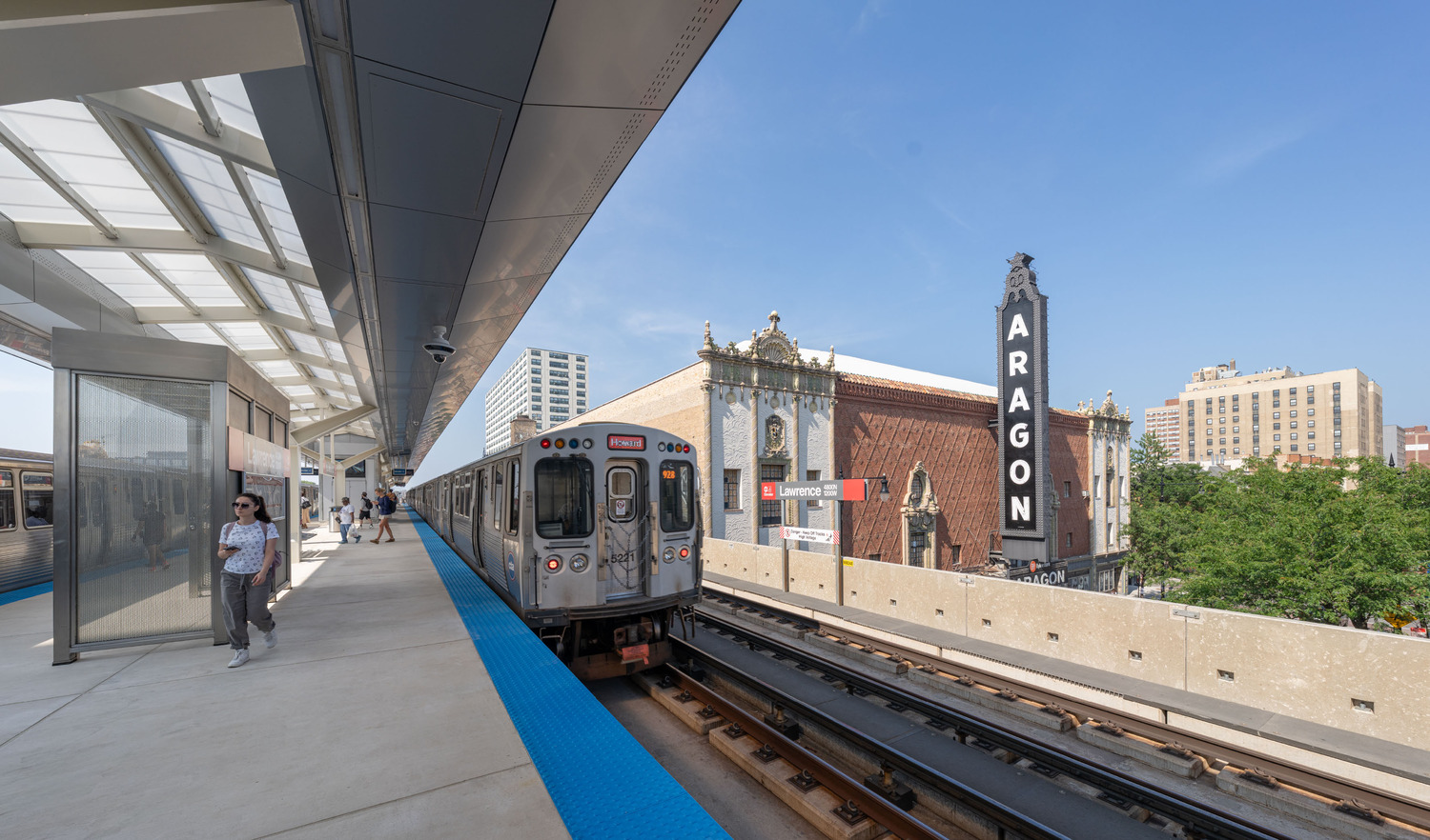 A train stopped at an elevated city station beside the Aragon theater, with a few people walking on the platform under a clear sky.