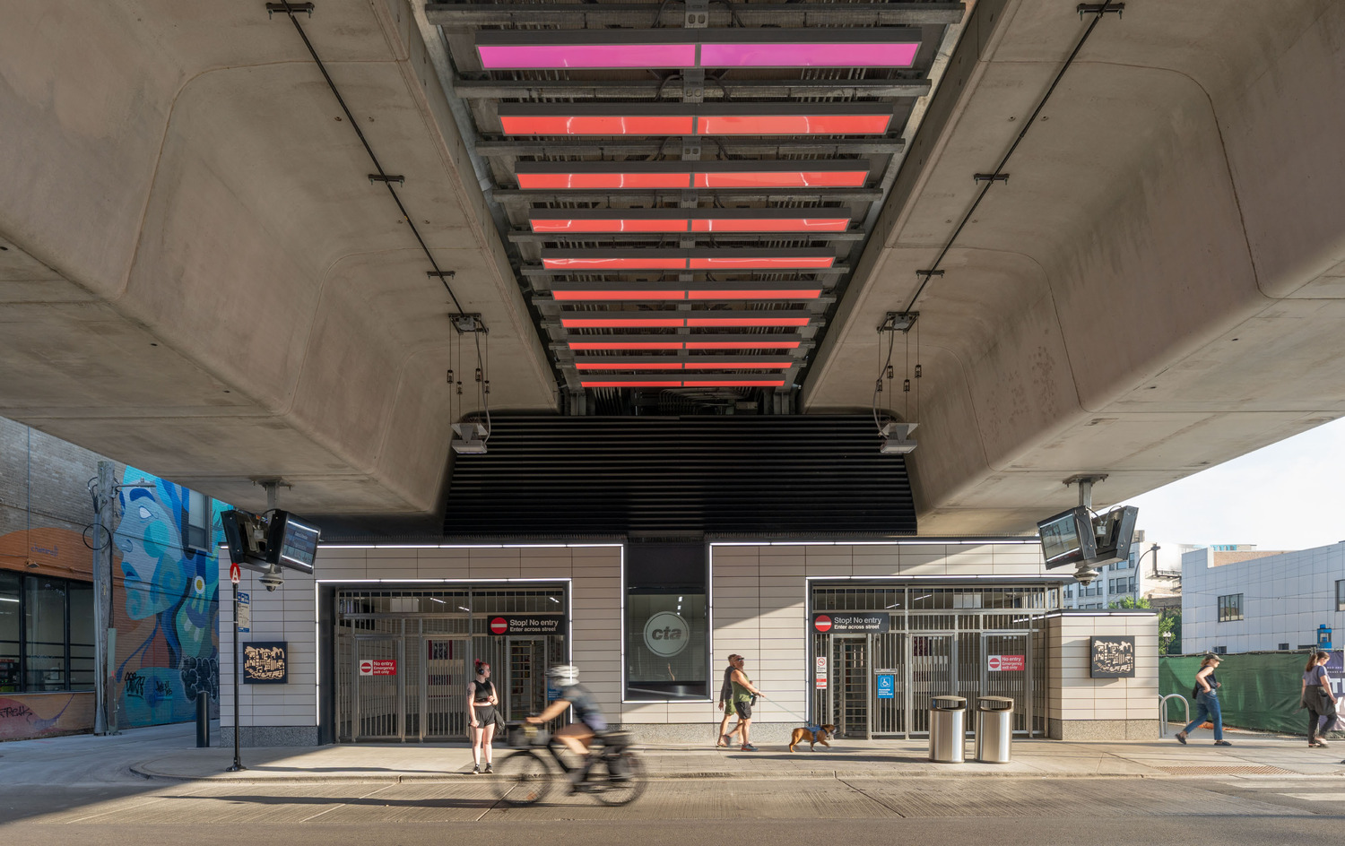 People walk and bike under an elevated train track with red lights above, near the entrance to a CTA transit station.