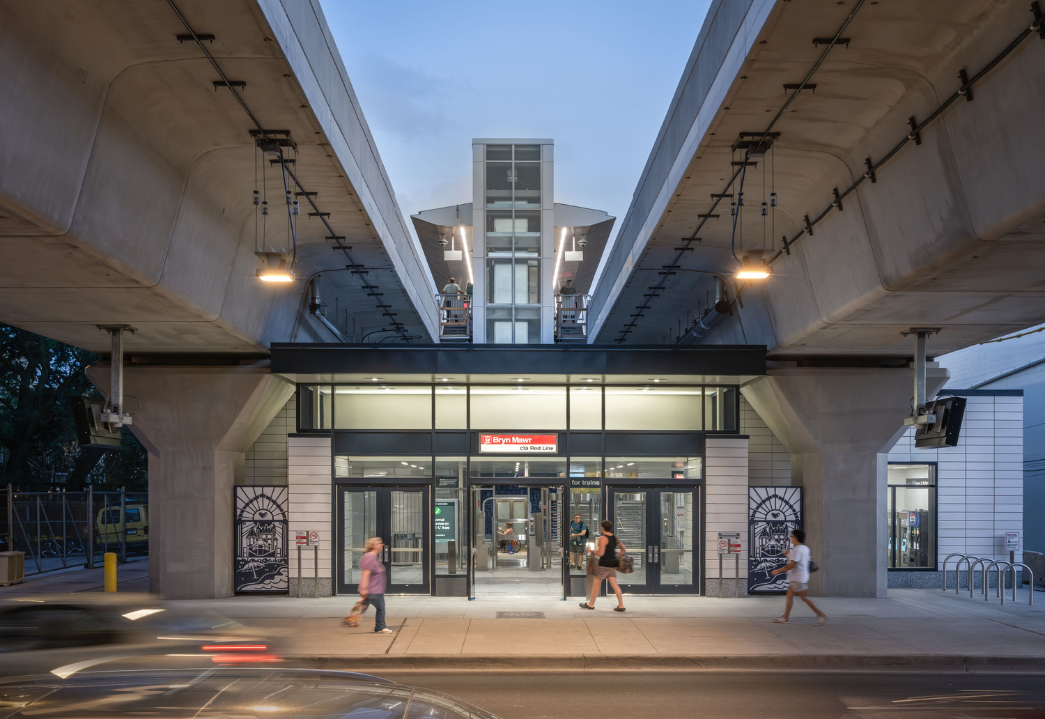 People walk past the entrance of a modern elevated train station at dusk, with staircases and platforms visible above.