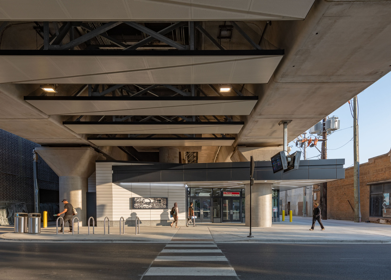A modern transit station entrance is beneath an overpass, with bike racks, pedestrians, and a digital display visible near the entrance.