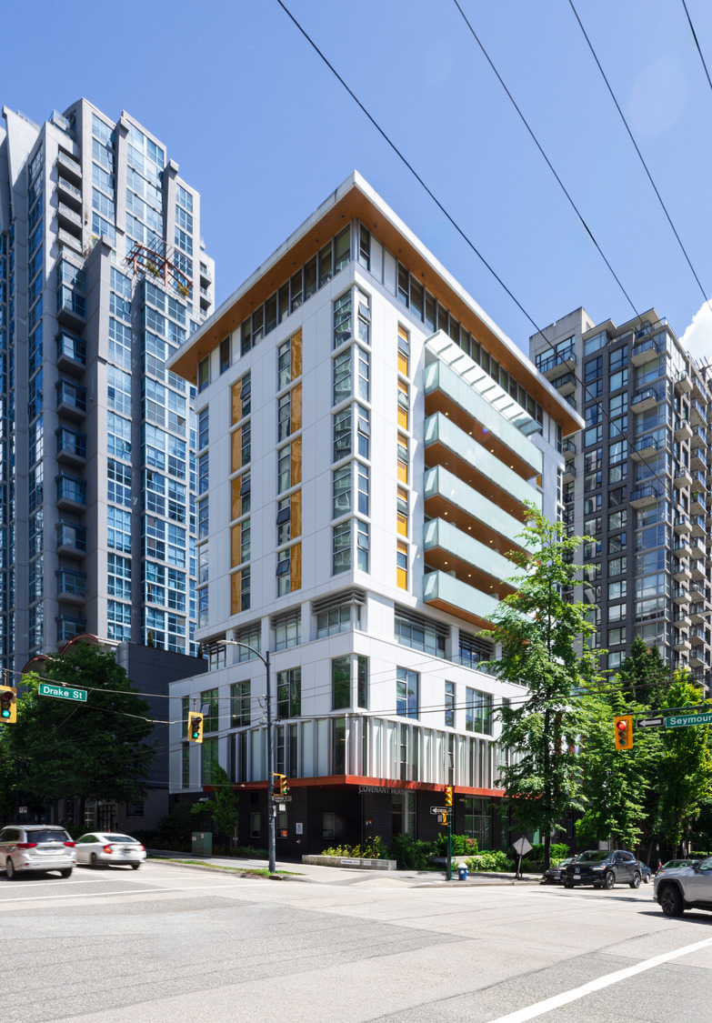 Modern mid-rise apartment building on a city corner, surrounded by taller glass buildings, with cars and traffic lights at the intersection.