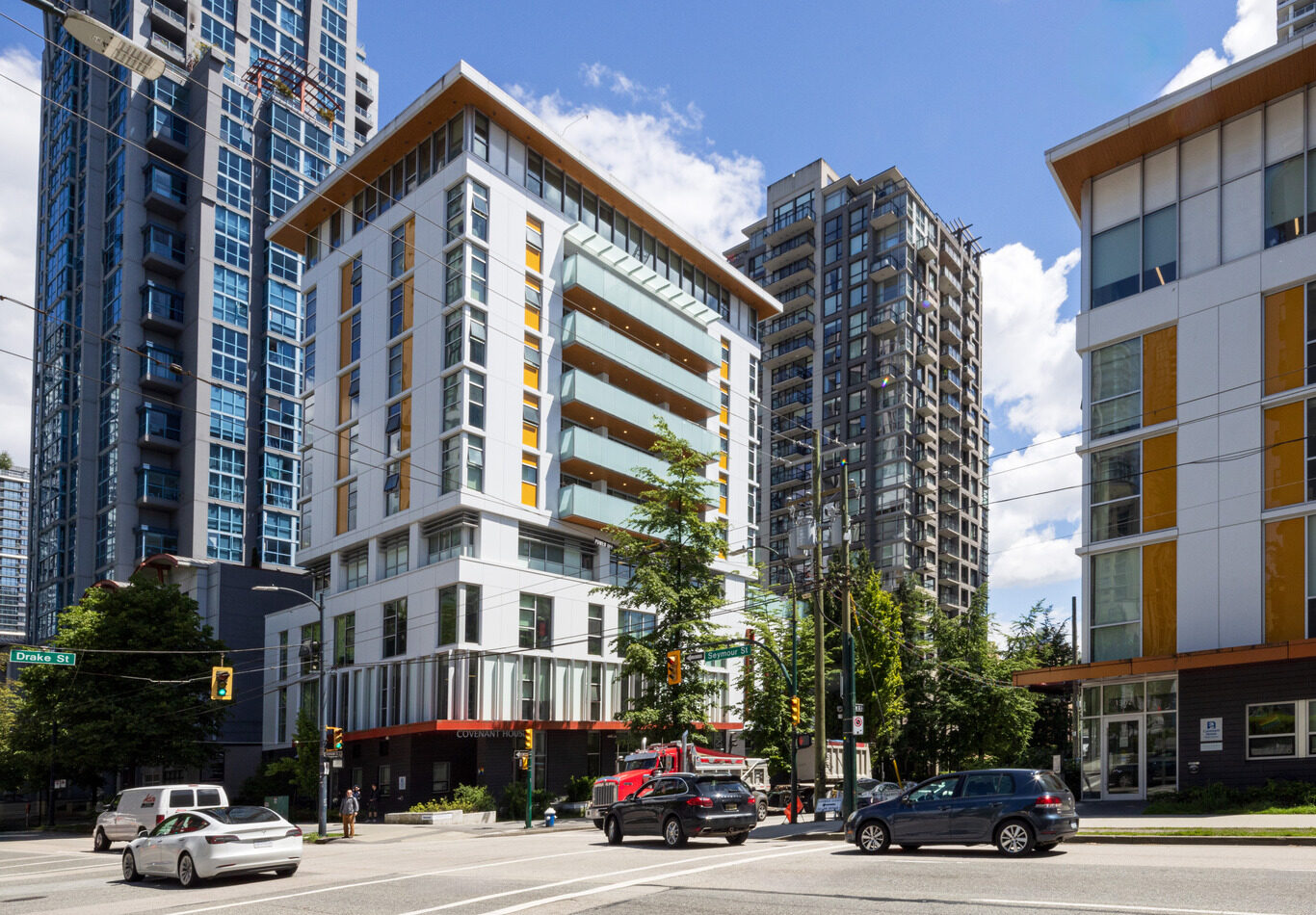 A city intersection with modern mid-rise and high-rise buildings, cars waiting at a traffic light, and a clear blue sky with some clouds.