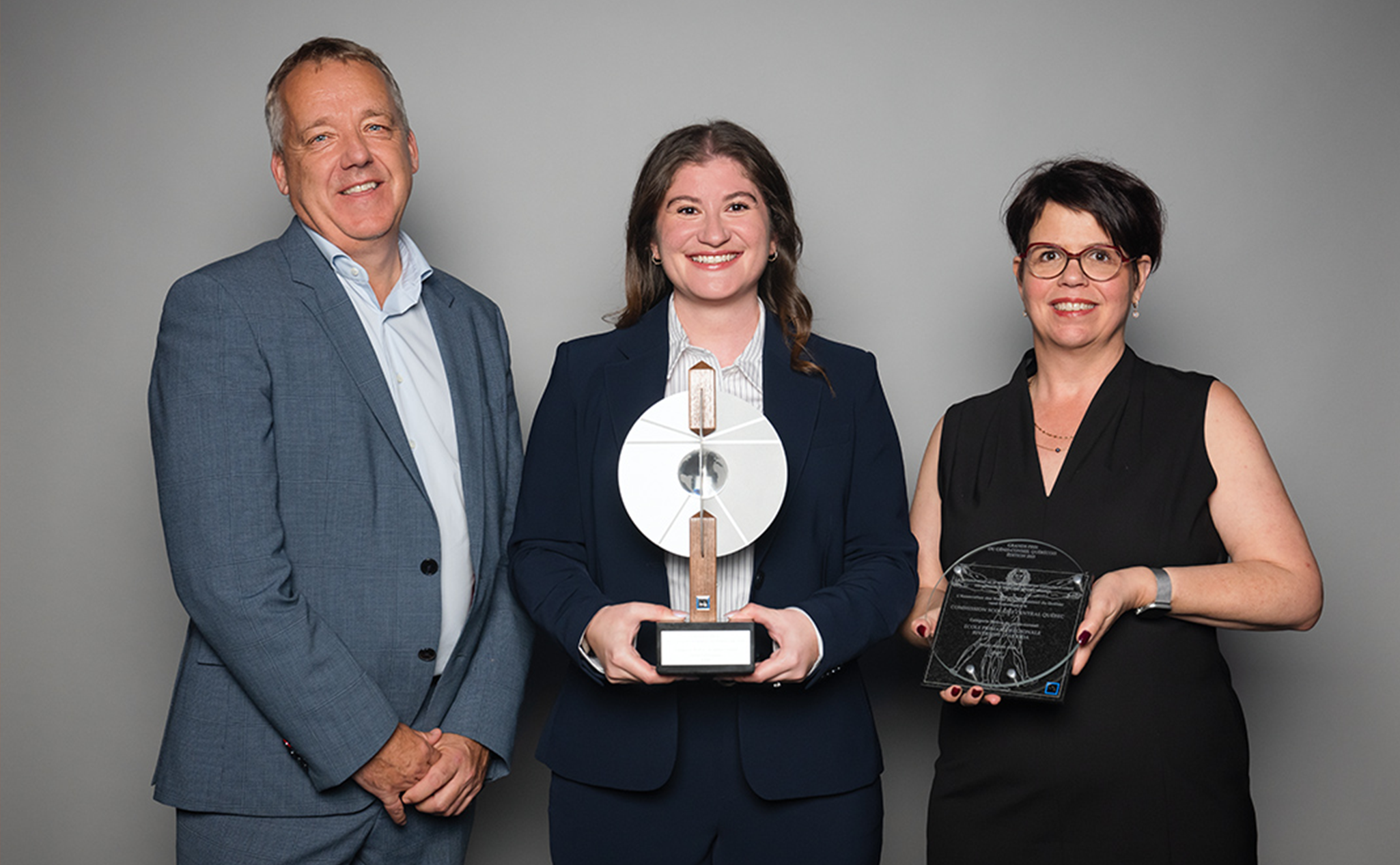 Trois personnes posent sur un fond gris ; la femme au milieu tient un grand trophée, et la femme à droite tient une récompense en verre.