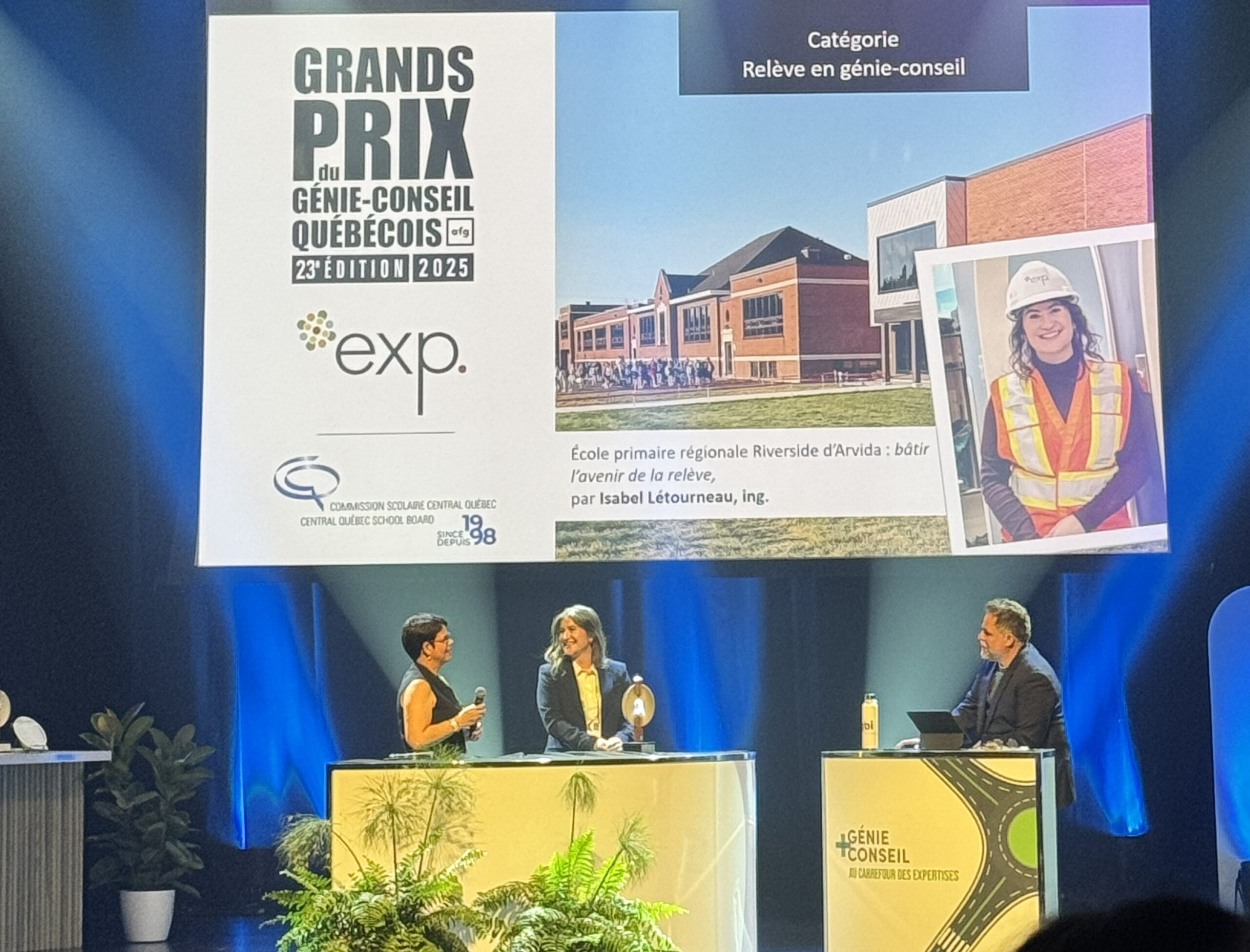 Trois personnes sont sur scène lors d'une cérémonie de remise de prix. Un grand écran affiche les détails du projet, une photo du bâtiment et une femme portant un gilet de sécurité et un casque. Il s'agit d'un concours d'ingénierie au Québec.
