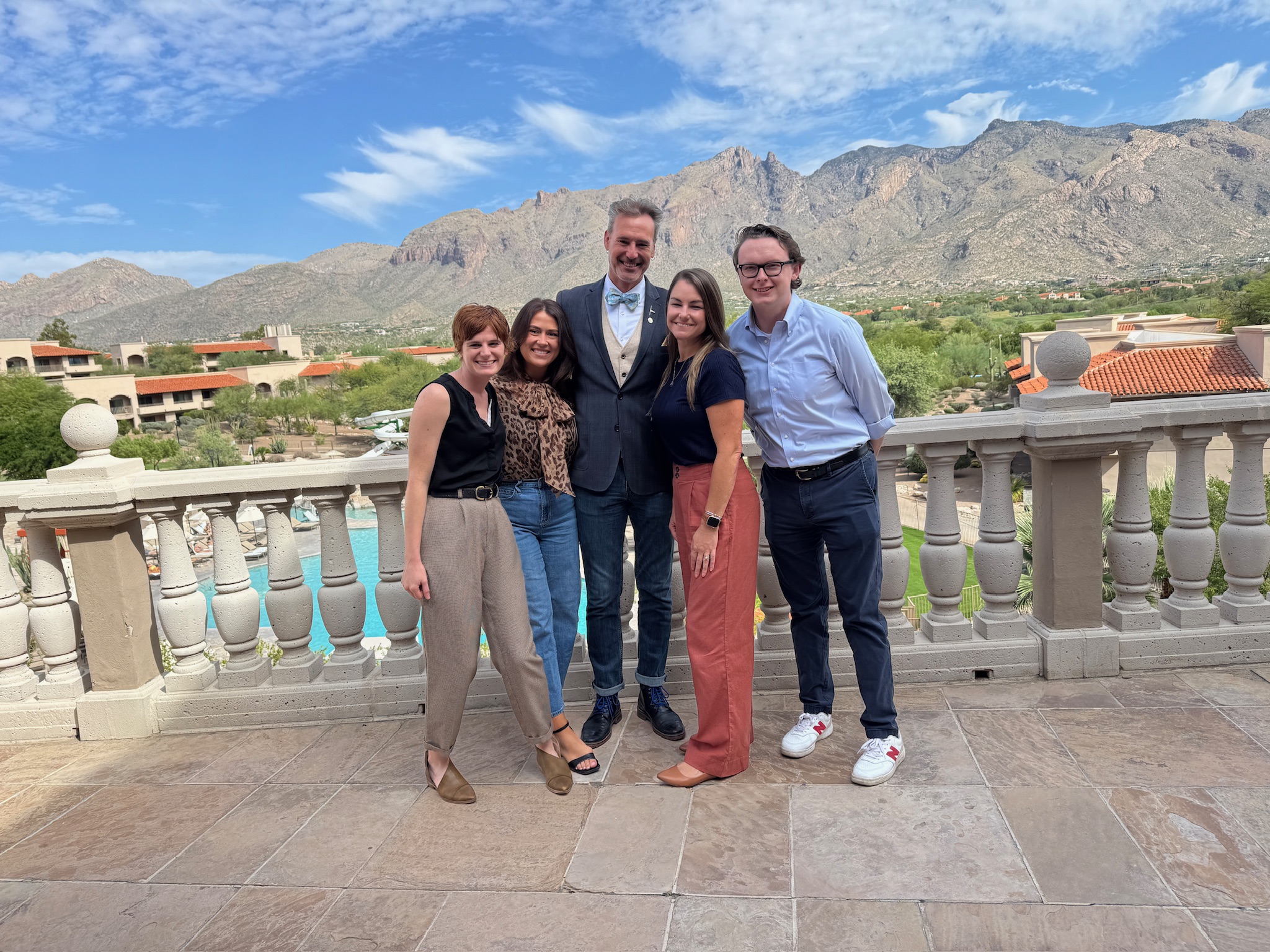 Five people pose together on a stone terrace with mountains, blue sky, and buildings visible in the background.