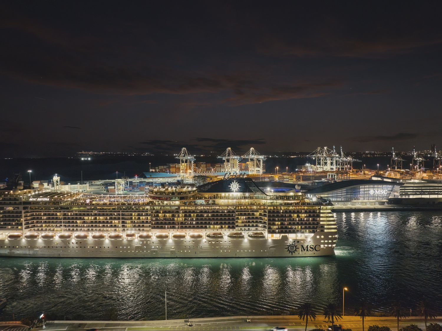 Two large MSC cruise ships are docked at a brightly lit port at night, with cranes and other ships visible in the background.