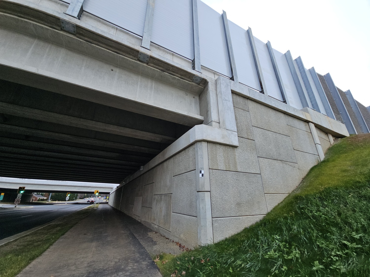 A concrete overpass with a sound barrier wall, a road and sidewalk passing underneath, and a grassy slope on the right side.