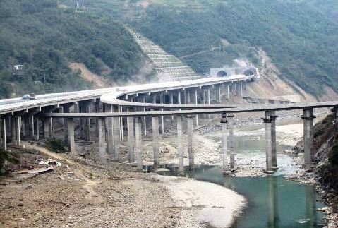 A multi-lane elevated highway curves across a river and valley, supported by tall concrete pillars, with tunnels visible in the hillside background.