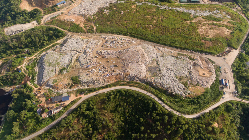 Aerial view of a large landfill surrounded by greenery, roads, and scattered structures on the edge of the waste area.
